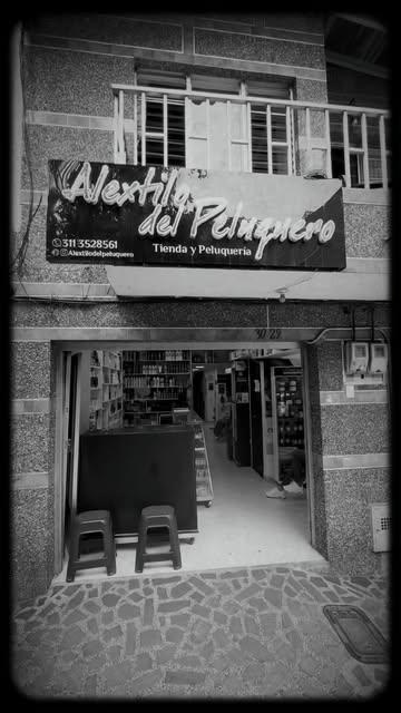 Foto en blanco y negro de la fachada: el letrero Alextilo del Peluquero sobre la puerta de garaje, con dos butacos en la entrada
